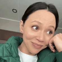 a woman wearing a green jacket and a white shirt has her hand on her chin