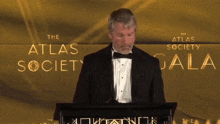 a man in a tuxedo stands at a podium in front of a wall that says the atlas society gala